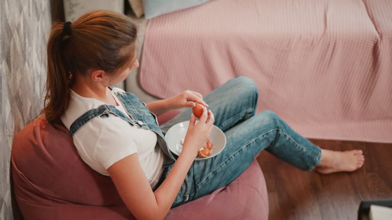Overhead view of woman seated on bean bag peeling egg into plate on her lap, casual indoor setting with soft lighting and cozy background textures