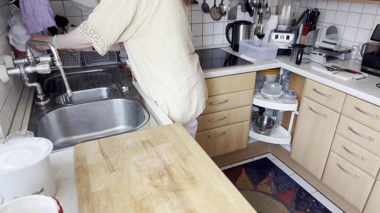 Woman wets a dishwashing brush and applies dish soap after organizing the kitchen, preparing to wash dishes at the sink. Static Medium Shot.