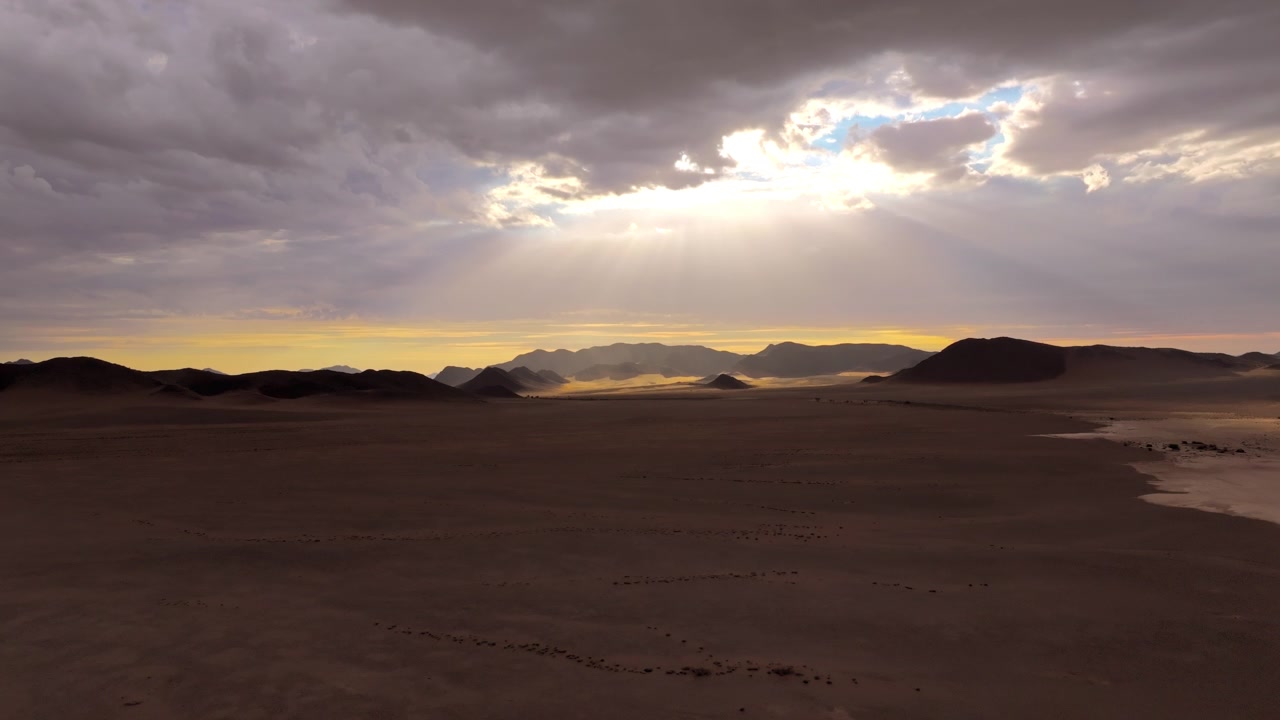 Ripresa aerea cinematografica di raggi di sole che filtrano attraverso nuvole pesanti sulle dune del deserto del Namib. Luce soffusa e nebbia che si sposta avvolgono le antiche dune e montagne in un'atmosfera cinematografica calma