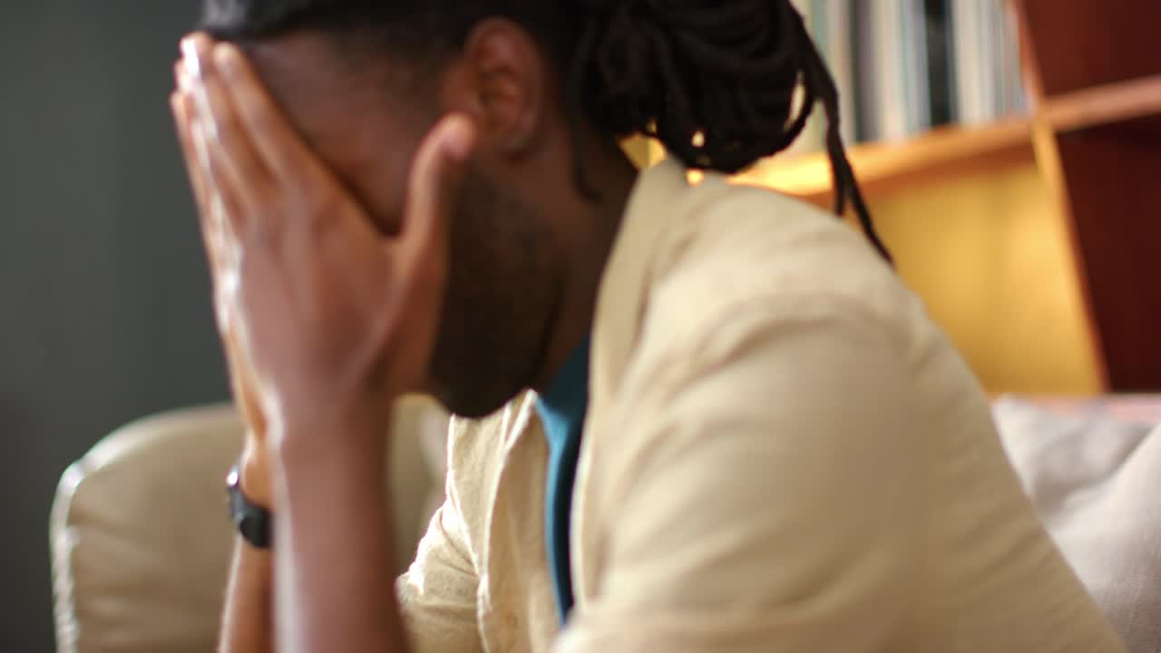 Un uomo africano si china in avanti sul divano, con le mani alla bocca che gli coprono il viso, strofinandolo e rivelando il profilo, pensieroso.