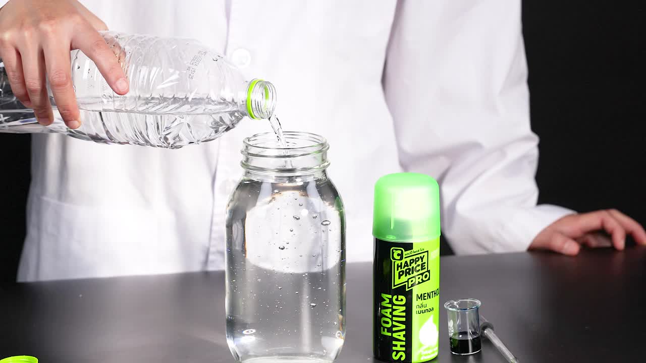 A person in a lab coat pours water into a jar, preparing for a chemical reaction experiment with shaving cream and dye