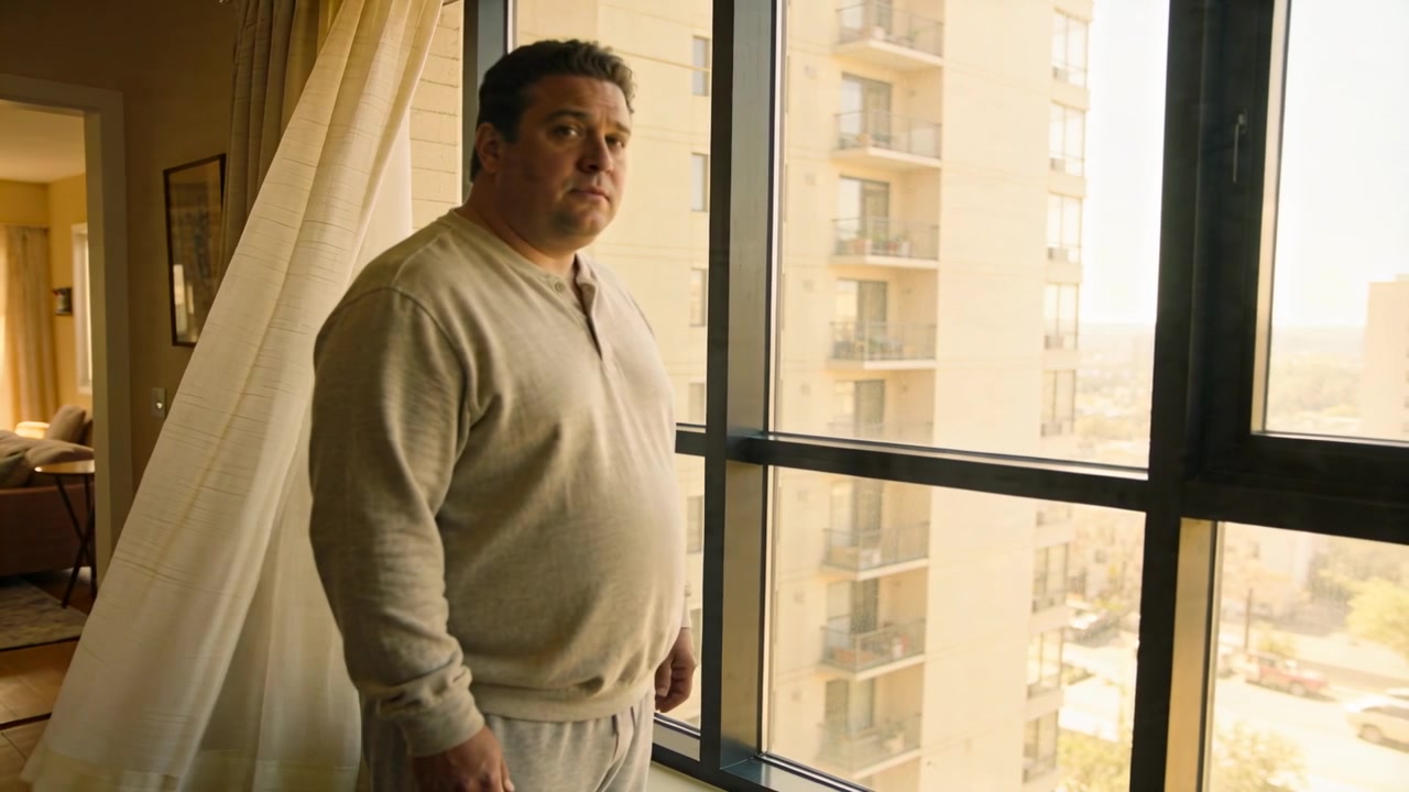 Hombre de pie con camiseta henley y pantalones de chándal en la ventana de una torre, con la cortina ondeando, observando los balcones y la calle.