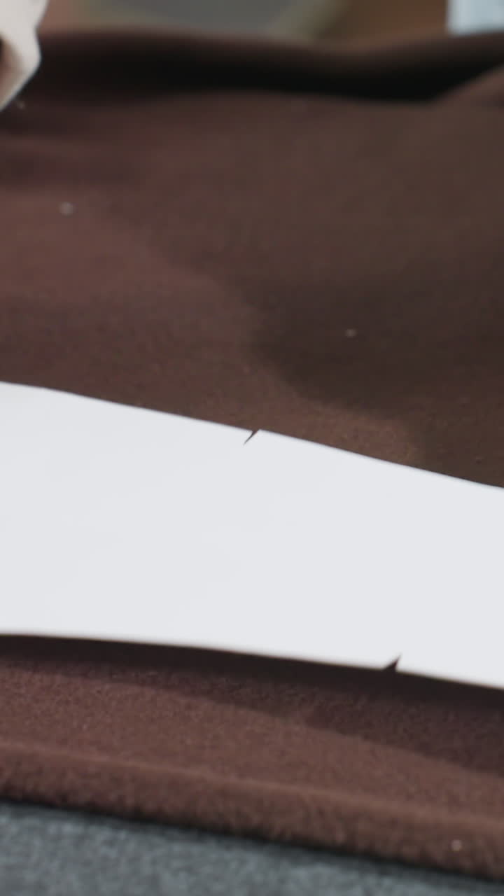 vellón marrón sobre la mesa, manos caucásicas alisando papel de patrón, sastre casero alineando la plantilla blanca, inspección táctil de la tela, primer plano de textura suave, alisado preciso de los bordes, iluminación acogedora de estudio