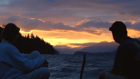 two people talking while watching vibrant sunset on a lake with mountains and clouds in the background