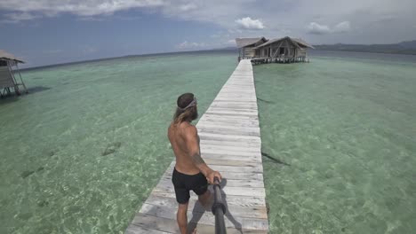 Un-Hombre-Joven,-Fuerte-Y-En-Forma-Con-El-Pelo-Largo-Y-La-Barba-Camina-Por-El-Embarcadero-De-Madera-Durante-Un-Día-Soleado-Y-Sonríe-A-La-Cámara-Mientras-Se-Filma-A-Sí-Mismo-Caminando-A-Su-Bungalow-De-Lujo-Desde-La-Playa