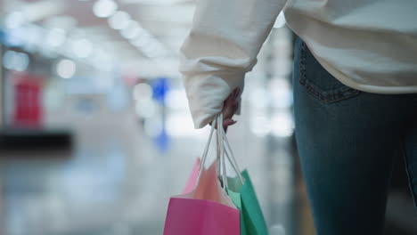 pink shopping bag held in someone's hand, accompanied by mint-colored bag, set against vibrant shopping mall background featuring colorful reflections, modern retail displays