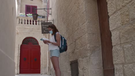 female tourist in facemask wandering in the steert of mdina looking for destinatination through guidebook in malta