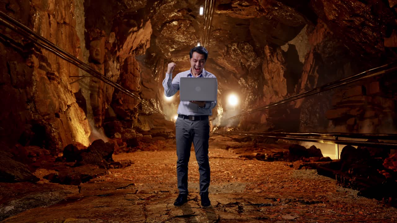 Full Body Of An Asian Male Professional Worker Standing With His Laptop In Underground Mine Tunnel, He Raises His Fist Up With Screaming Goal