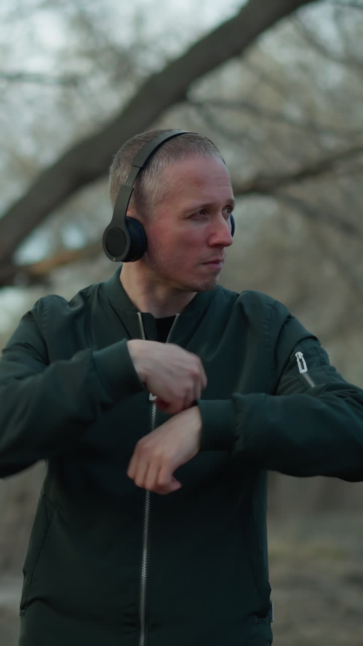 A man in a green jacket and headphones dances with expressive movements in a wooded area