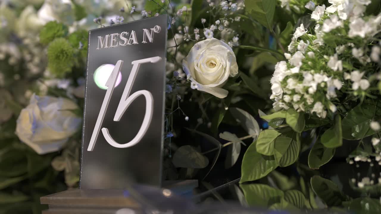 A close-up of an elegant wedding table centerpiece, featuring a modern black and white "MESA Nº 15" sign surrounded by lush white roses and greenery