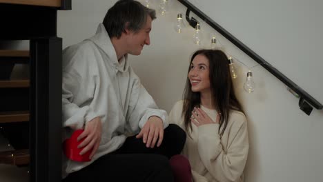 smiling woman receiving a gift from her husband for valentine's day while sitting on the stairs