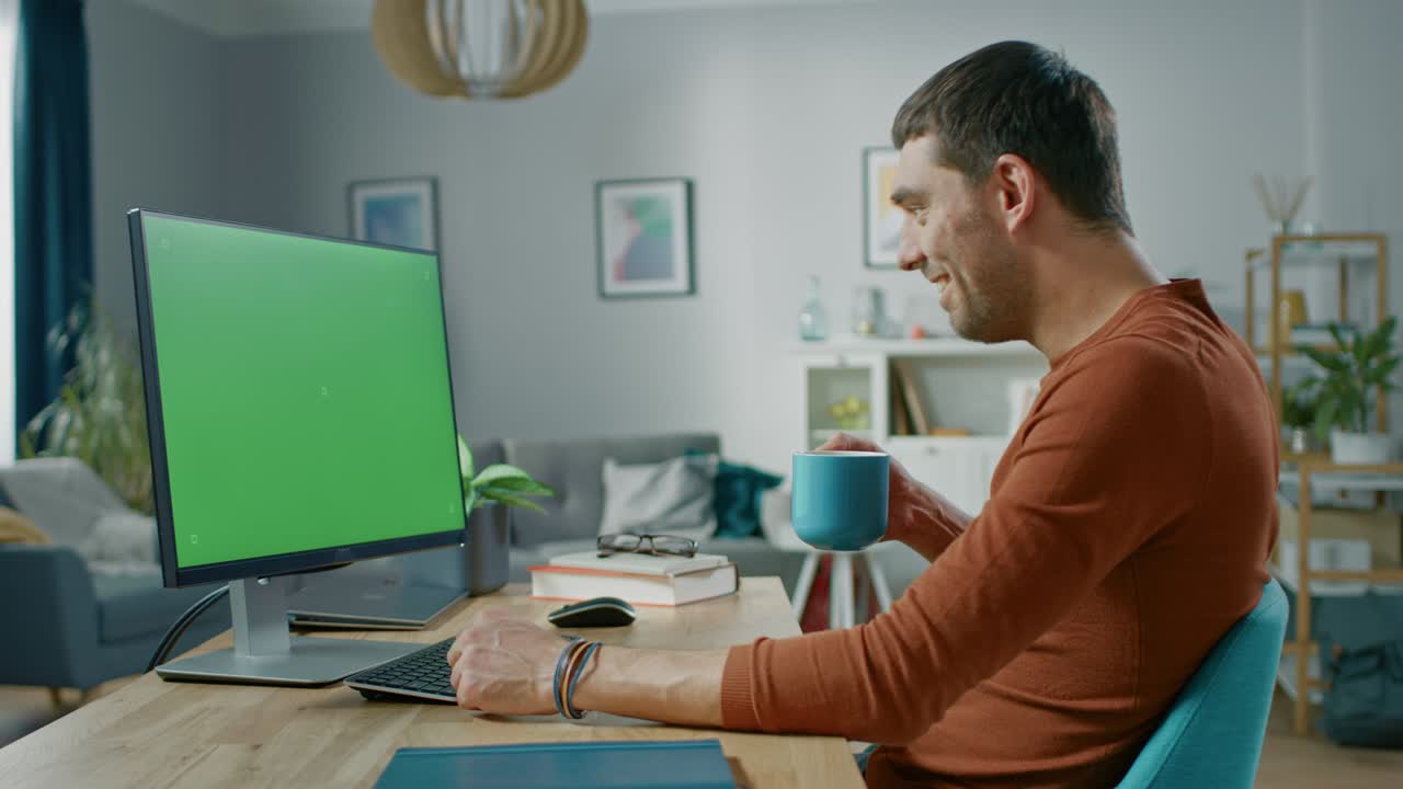 un bel uomo sorridente seduto alla sua scrivania a casa usa un personal computer con un modello di schermo verde. vive in un accogliente appartamento con un moderno interno in salotto.