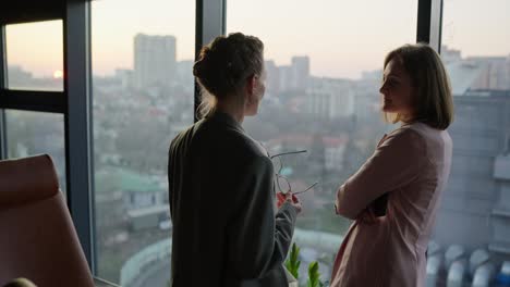 two middle-aged businesswomen in business uniforms stand near panoramic windows in a modern office and communicate about current issues