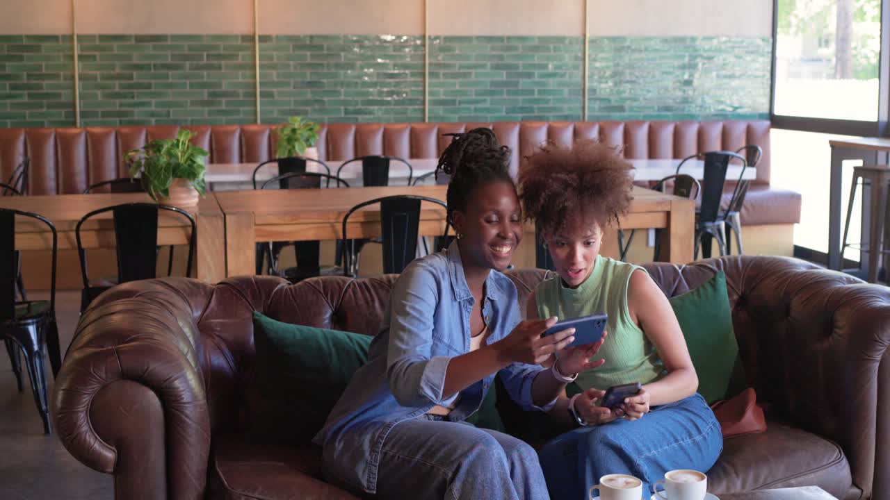 African American women friends cafe sofa seeing server tapping wrist sharing phones and taking sel