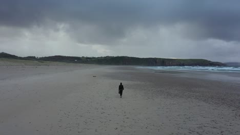 back view of man walking alone on empty deserted beach on winter cloudy day