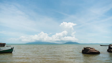 Meerblick-Zeitraffer-Einer-Insel-Am-Meereshorizont-Mit-Wolkenformationen-Am-Blauen-Himmel-über-Den-Hügeln-Mit-Fischerbooten,-Die-In-Die-Szene-Ein--Und-Ausfahren,-Felsen-Im-Vordergrund