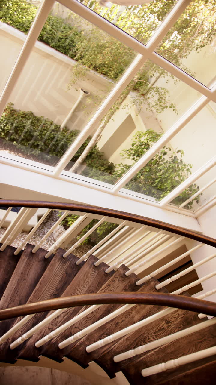 Vertical video: Curved wooden staircase with large windows overlooking lush green garden