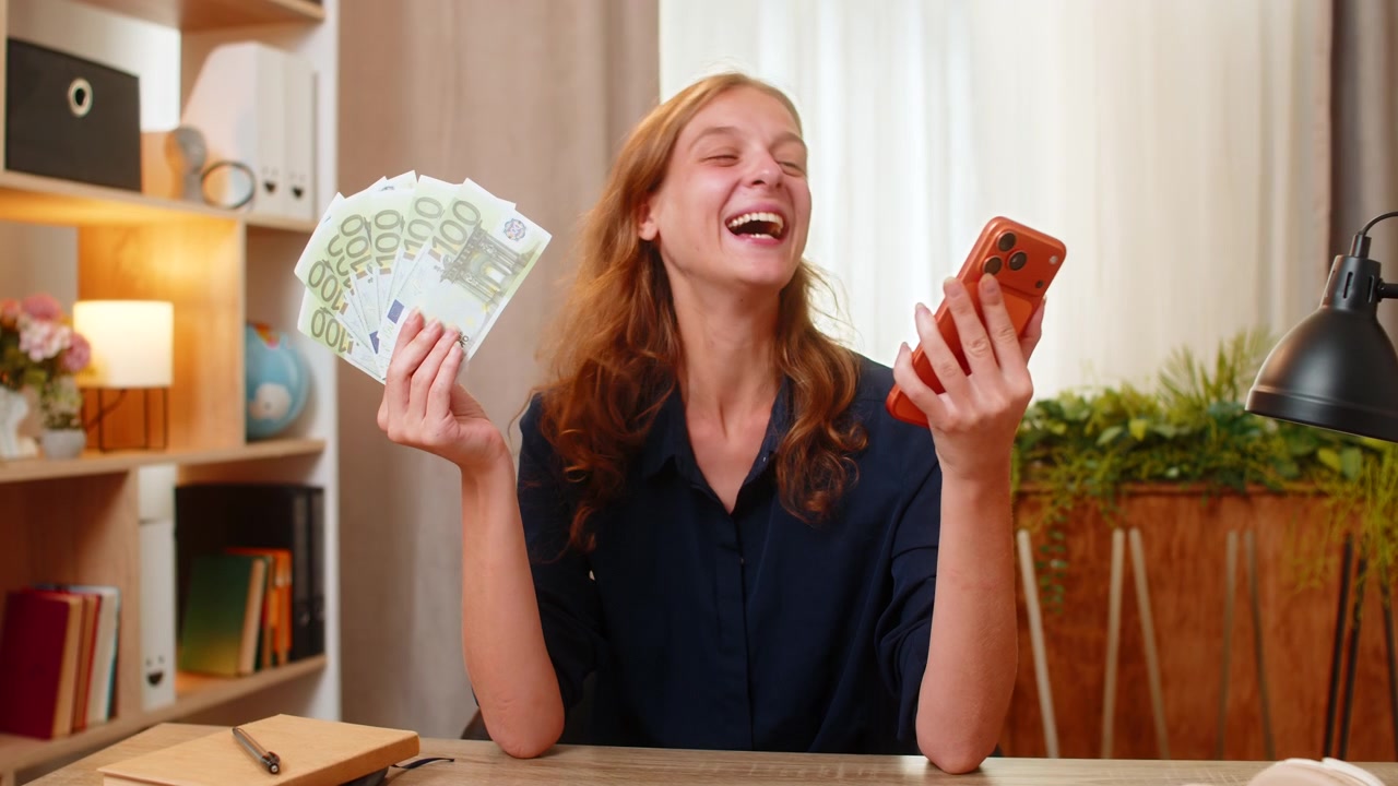 Jonge vrouw aan thuistafel houdt contant geld en smartphone vast, lachend na succesvolle online betaling thuis.
