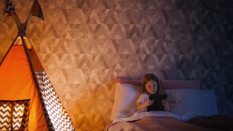 girl watches video via tablet in bed near play teepee