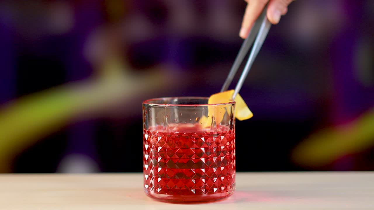 Un cocktail Negroni viene preparato con una guarnizione di scorza d'arancia in un ambiente vivace e colorato.