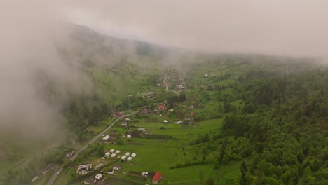 Aerial-descending-from-clouds,-revealing-a-small-rural-city-nestled-in-a-valley-between-mountains-surrounded-by-green-forests