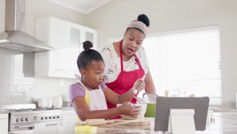 happy unaltered african american mother and daughter baking in kitchen using tablet, in slow motion