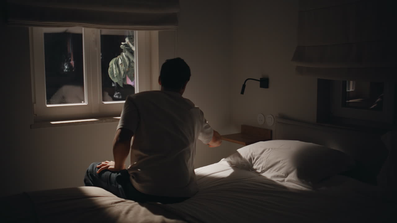 Troubled man sitting night bedroom home by window feeling melancholic back view