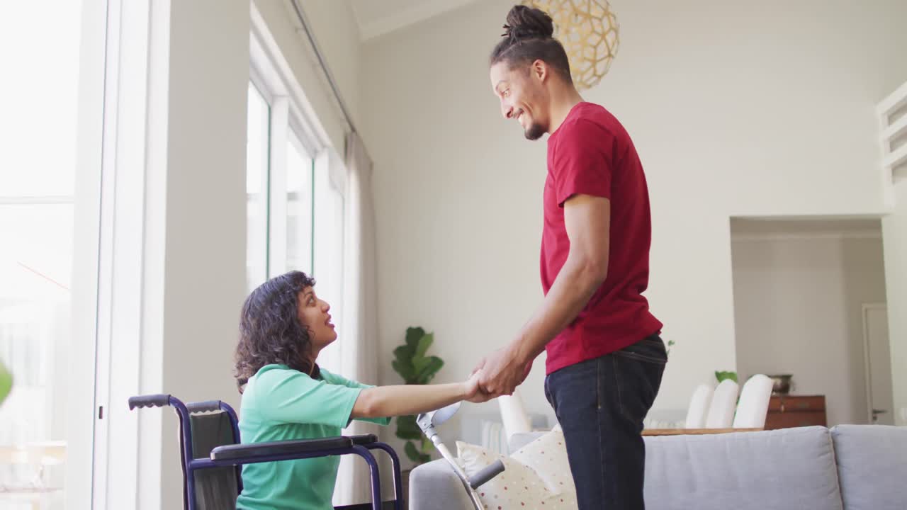 mujer biracial feliz en silla de ruedas y compañero masculino sonriente tomándose de la mano y hablando en la sala de estar