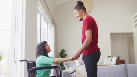 Happy-biracial-woman-in-wheelchair-and-smiling-male-partner-holding-hands-and-talking-in-living-room