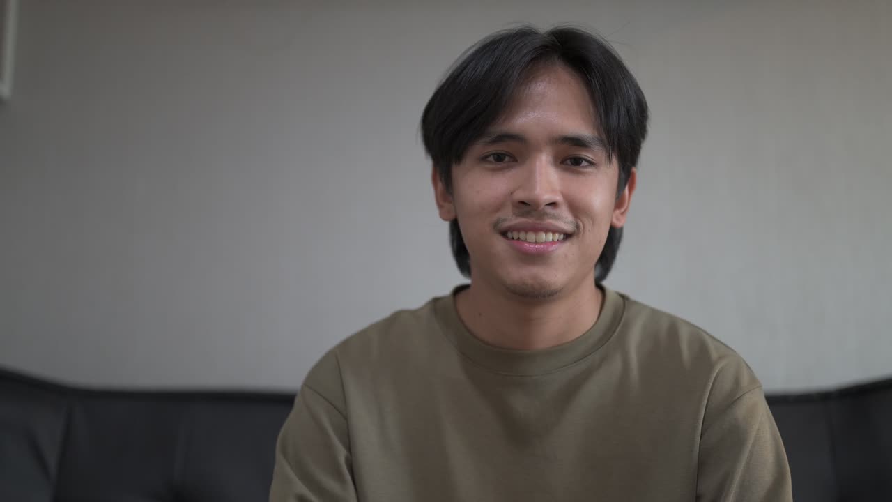 Happy young Asian Thai man in early 20s sitting at sofa indoors while vlogging and talking online