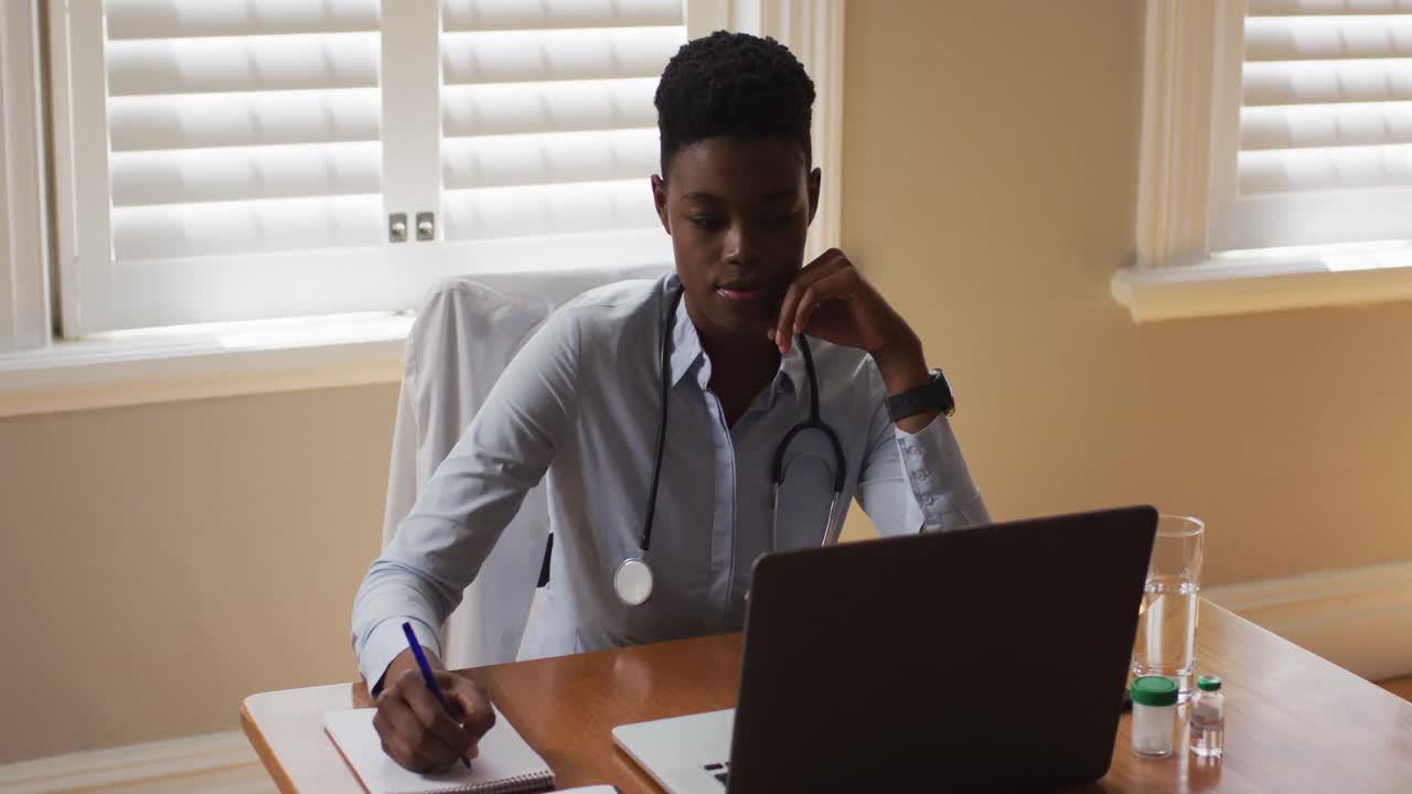 African american female doctor taking notes while having a videocall on laptop at home