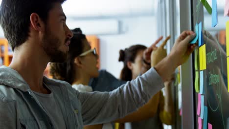 Male-executive-writing-on-sticky-notes-while-colleagues-discussing-during-meeting