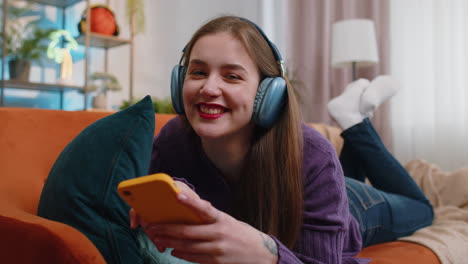 happy young woman in wireless headphones relaxing lying on sofa at home listening favorite music