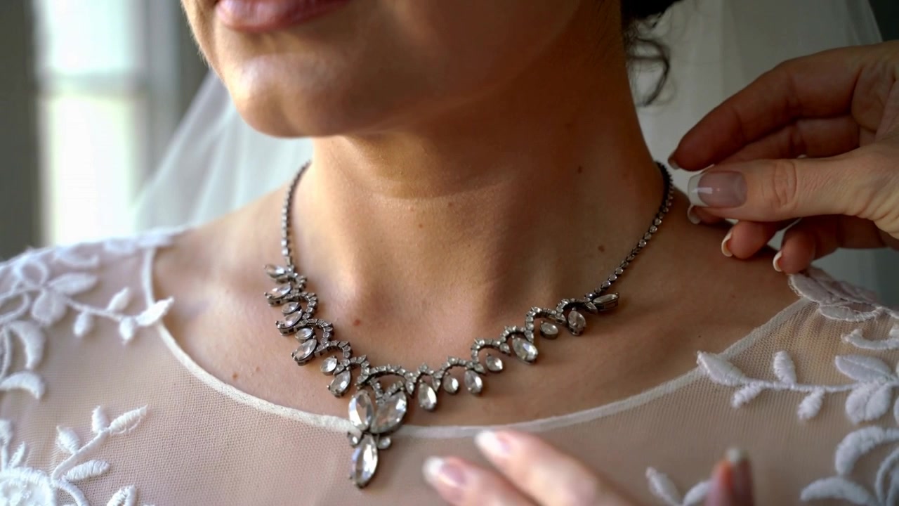 Un'assistente tiene una collana e allaccia la chiusura alla scollatura della sposa nella suite nuziale, con il velo già indossato per la cerimonia.