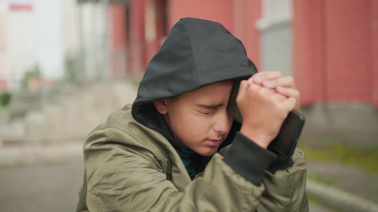 Teen in hooded jacket seated outdoors holding phone against forehead with eyes closed tightly, then opening eyes slowly while appearing deep in thought near brick wall and pavement