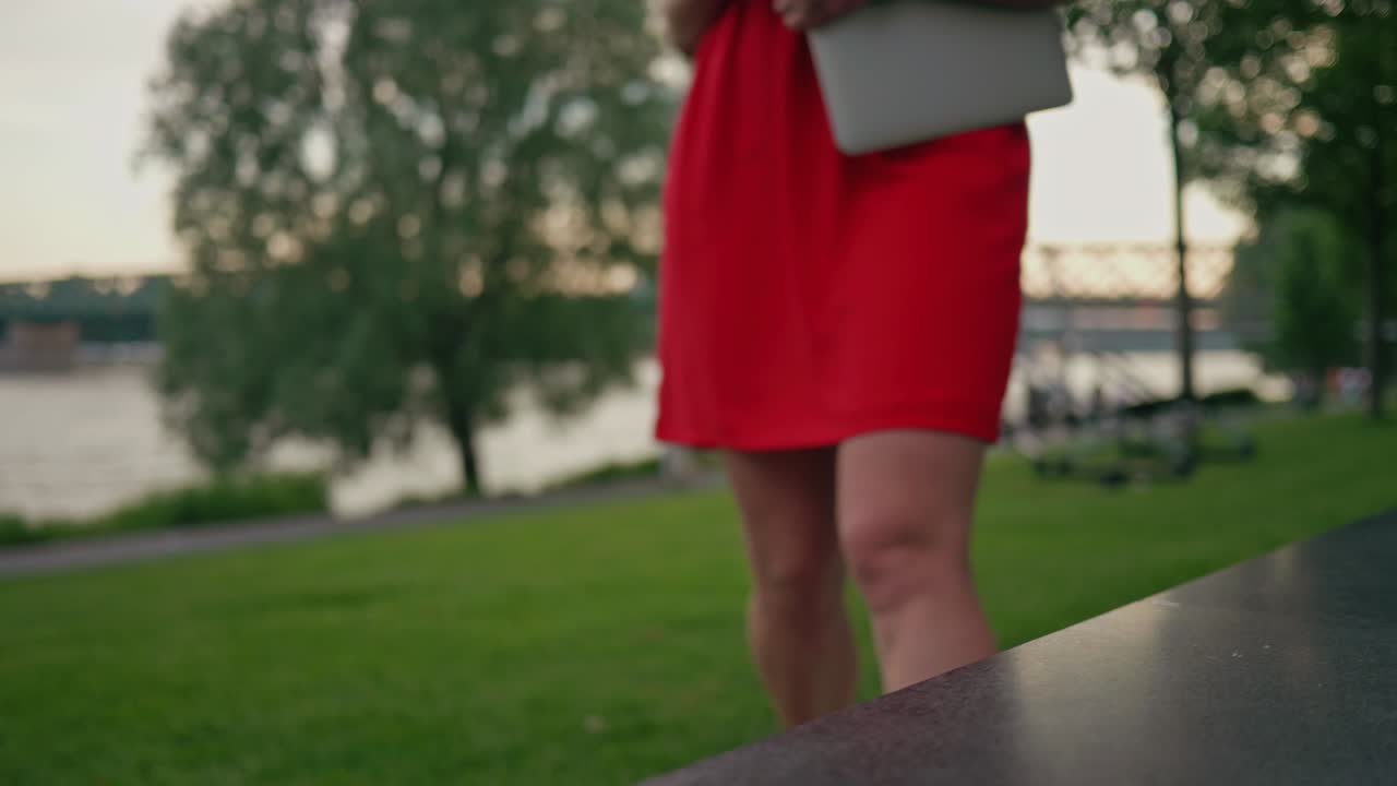 hermosa mujer vestida de rojo con una laptop caminando hacia el banco en el parque y sentada en ella