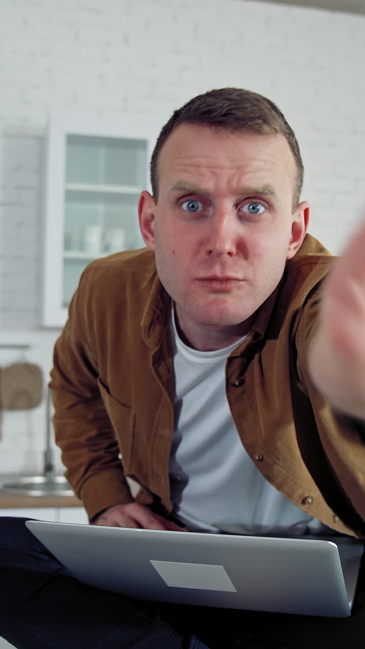Funny comic businessman working at home. Mad face of a young freelancer looking on camera angrily while sitting on a kitchen table with a laptop. Vertical video