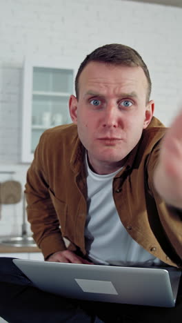 Funny comic businessman working at home. Mad face of a young freelancer looking on camera angrily while sitting on a kitchen table with a laptop. Vertical video