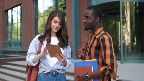 caucasian woman and african american man talking and watching something on the tablet in the street near the university