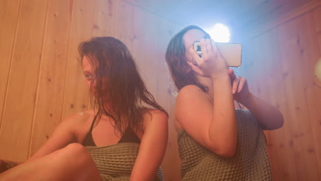 two women in towels sit on wooden sauna bench in smoky haze under warm colored lights while one holds smartphone taking mirror selfie capturing playful spa friendship