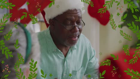 Animation-of-green-leaves-over-senior-african-american-man-in-christmas-hat-saying-grace-at-dinner