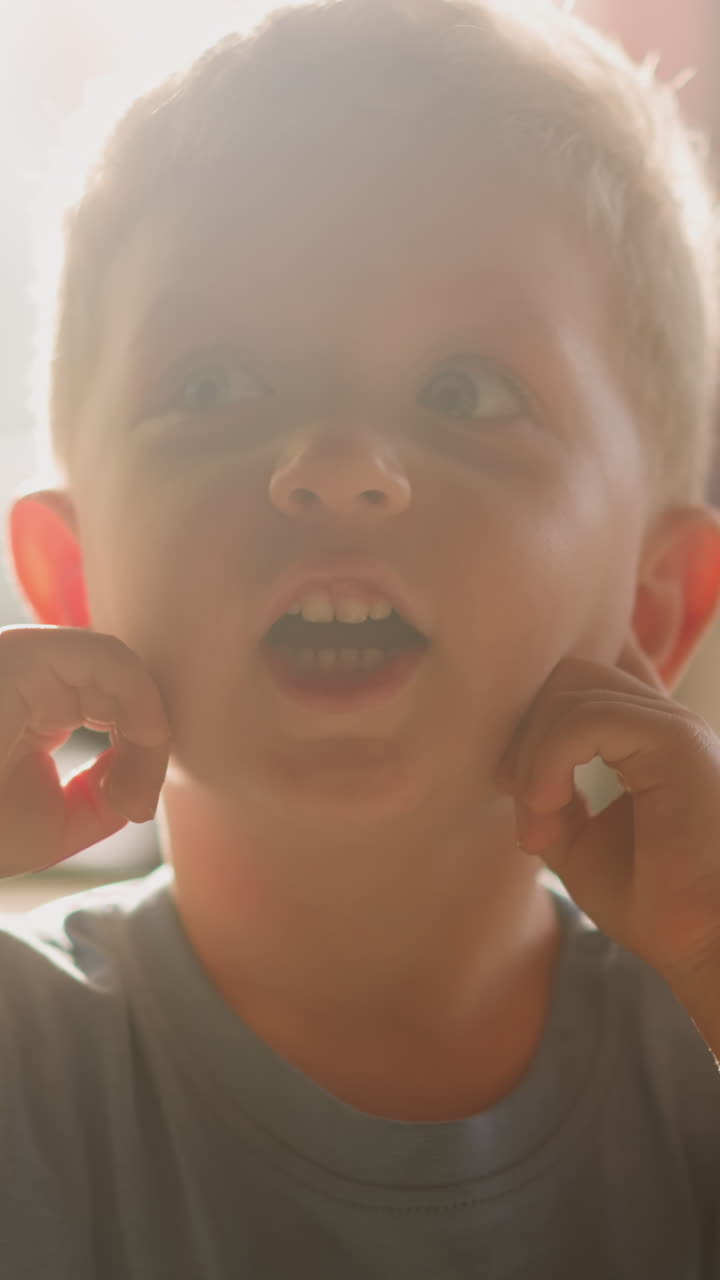 Playful little boy with open mouth closes ears by fingers having fun in room lit by bright sunlight at home close view slow motion