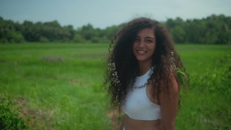 young woman with middle eastern features, long dark curly hair, and a white top turns smiling toward the camera in a green field, with small white flowers tucked over her shoulder in open landscape