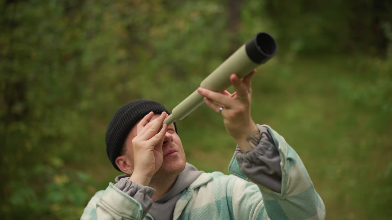 Astrofilo caucasico amatoriale che testa un telescopio portatile in una pineta verdeggiante, indossa un berretto di lana e una giacca a quadri, regola la lente e l'angolo con espressione curiosa e concentrata durante un esperimento diurno