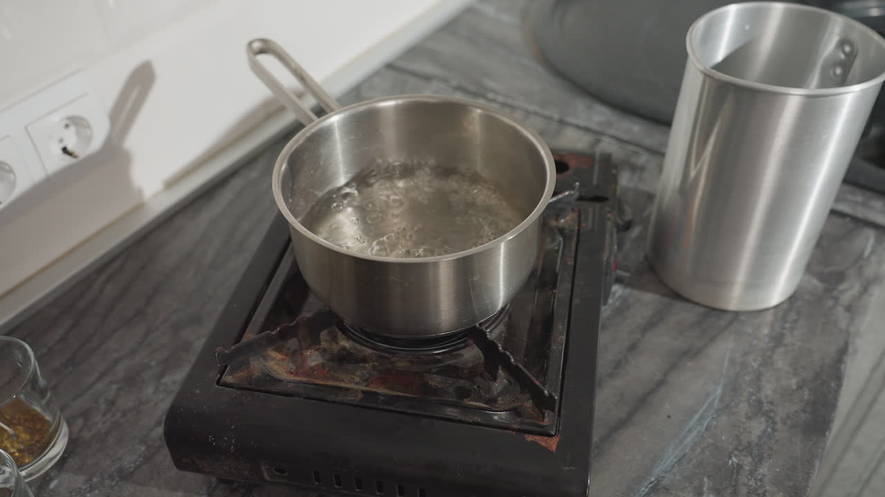 Boiling water steaming vigorously in stainless pot placed on gas burner, with silver jug standing beside pot on marble countertop and glass cups with candle wicks nearby, kitchen setting background
