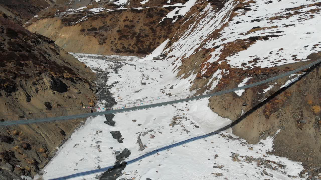 Wide Aerial shot of an empty suspension bridge with some Buddhist prayer flags over a frozen river (Marshyangdi) in the Annapurna circuit trek, Manang, Nepal