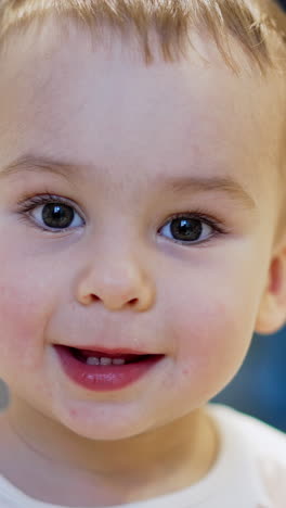 beautiful caucasian baby portrait. adorable toddler boy sits calm smiling to the camera and approaching it. close up. blurred backdrop. vertical video