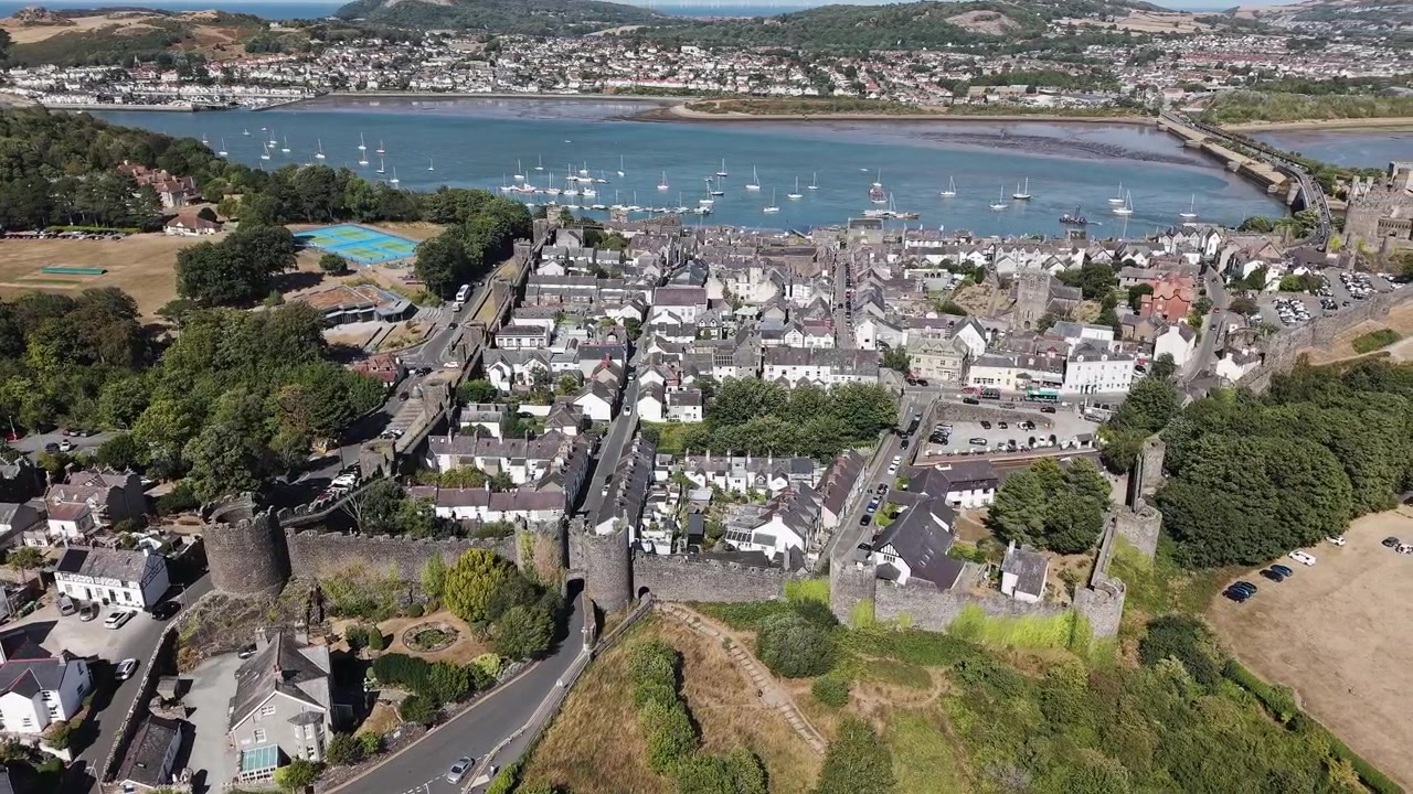 Medieval stone fortifications enclosing a coastal town in Wales, boats moored in calm estuary waters under clear sky