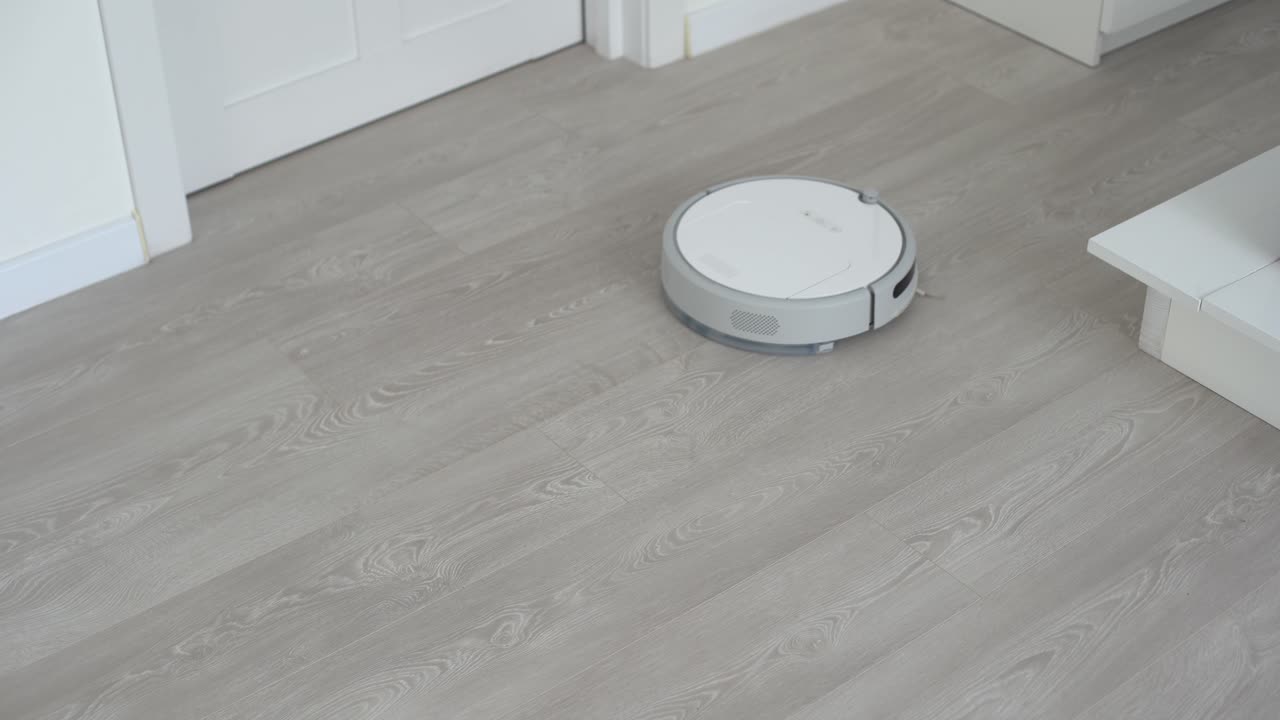 белый робот-пылесос на ламинированном полу очищает пыль в интерьере гостиной. умная технология уборки.