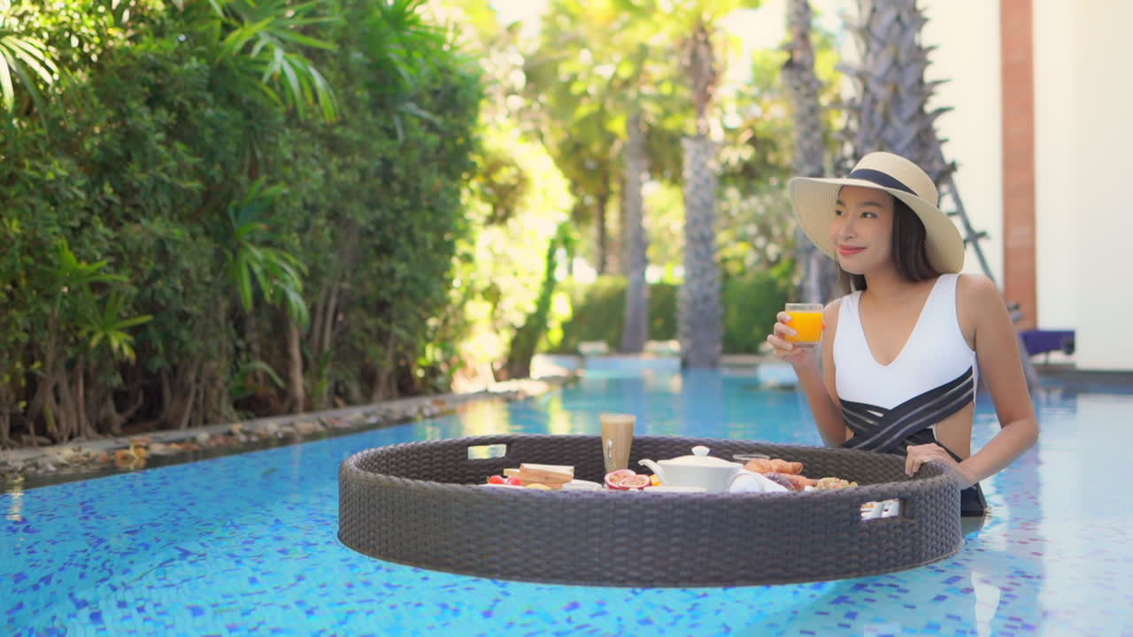 Attractive Asian Female Spinning Breakfast Floating Plate in Pool and Taking Orange Juice. Luxury Vacation Concept